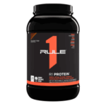 Envase de proteína en polvo R1 Protein de Rule 1, sabor chocolate fudge. Contiene 100% proteína de suero, con 25 gramos de proteína por porción y 28 porciones por envase. Ideal para apoyar el desarrollo muscular y la recuperación post-entrenamiento. Su fórmula incluye proteínas hidrolizadas para una absorción rápida y efectiva. Perfecto para deportistas y personas activas que buscan mejorar su rendimiento físico.