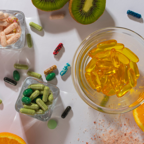 Imagen que muestra una variedad de suplementos nutricionales. En el centro, un recipiente transparente contiene cápsulas amarillas, rodeado de frascos con cápsulas de diferentes colores y formas. También se aprecian frutas como kiwi y naranja, que sugieren un enfoque saludable. La disposición de los suplementos y las frutas resalta la importancia de una dieta equilibrada y el uso de complementos para mejorar la salud. Ideal para contenido relacionado con nutrición y bienestar.